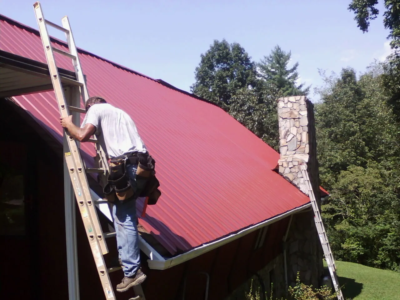Roofer on a ladder beside a red metal roof and stone chimney — Soffit & Fascia Repair by Roofing Brothers in Iuka